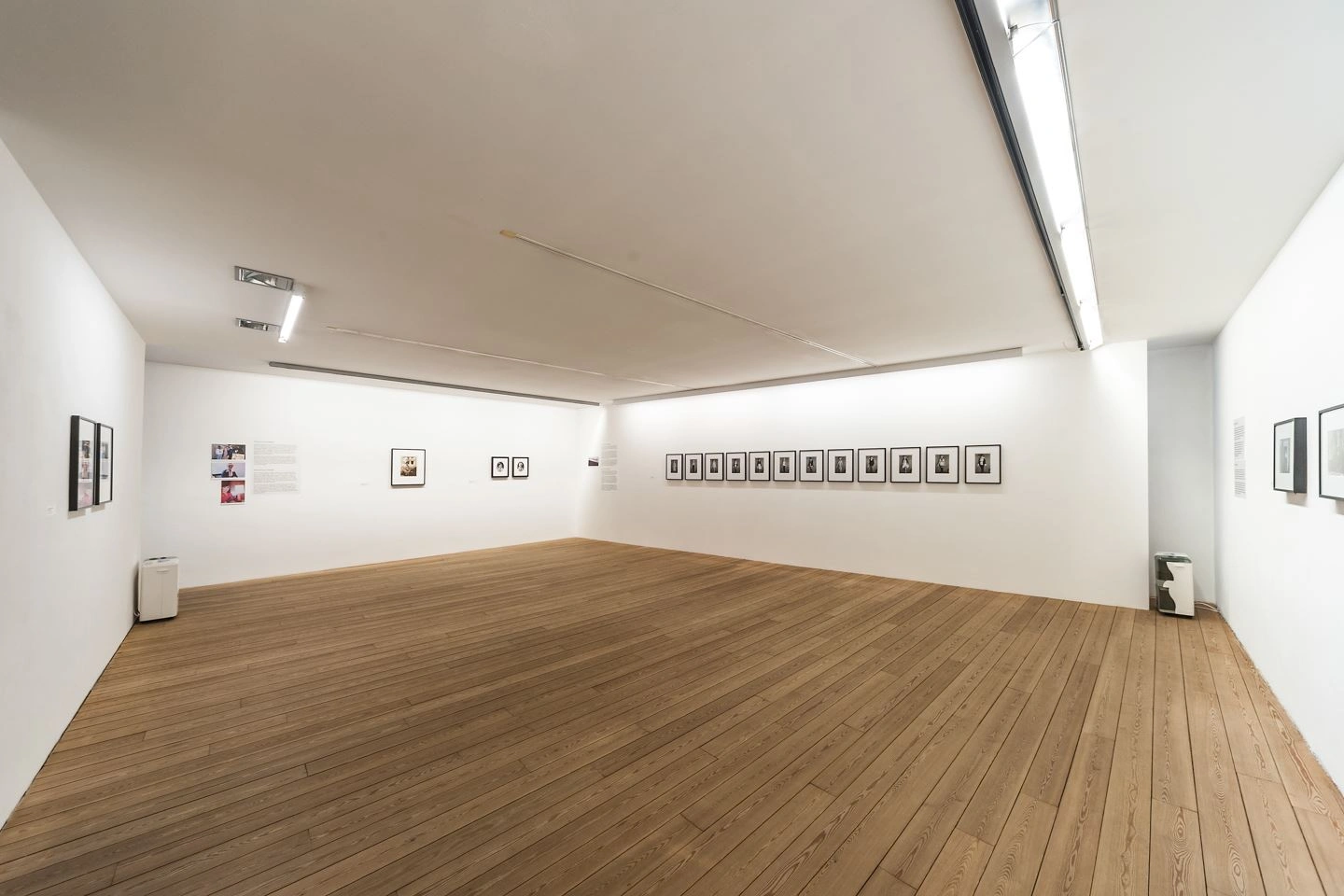 An exhibition space is shown on a large scale from the corner. Small works of art hang on each wall. There are two small objects in two corners.