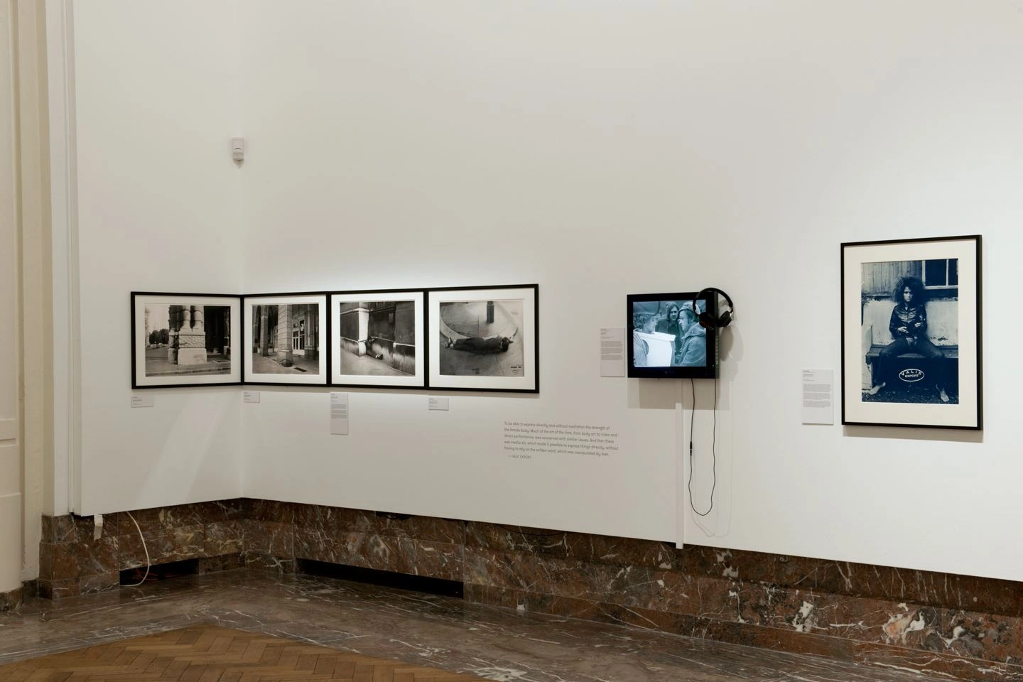 In einer Ausstellungshalle sind mehrere Kunstwerke von Bozar abgebildet. Links sind vier schwarz weiß Fotographien in Querformat aufgehängt, in der Mitte ein Monitor  mit Video und rechts eine Fotographie einer sitzenden Person in Hochformat.