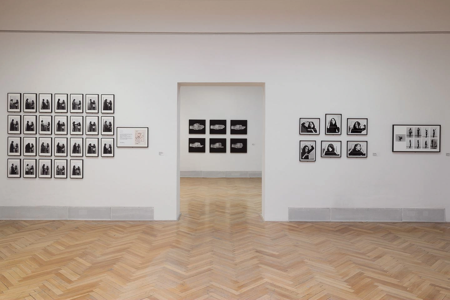 An exhibition wall in white with photographs on it can be seen. In the middle of the wall is a passageway to another exhibition room, where photographs also hang on the wall.