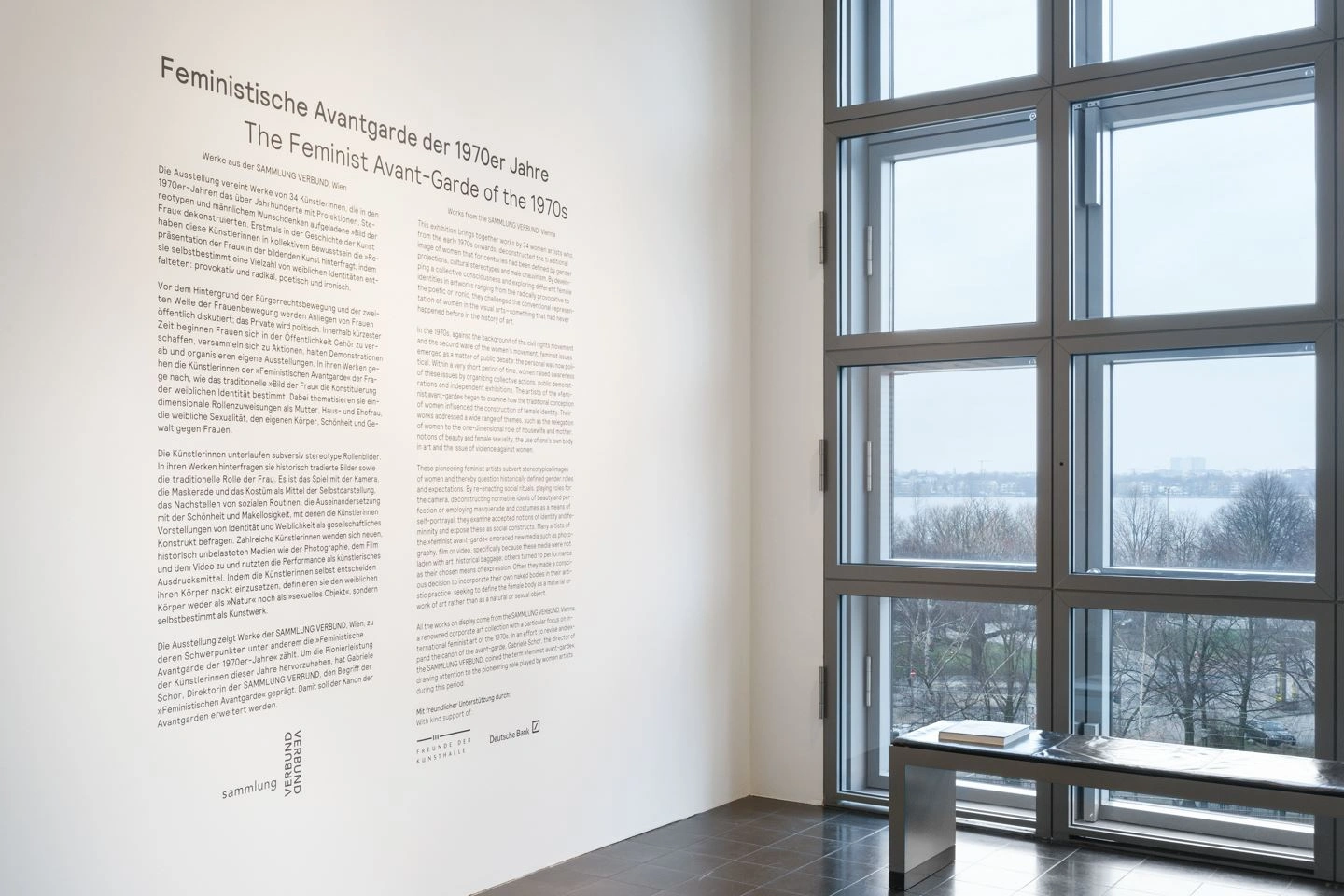 A modern room can be seen with a French window on the right. In front of it is a bench with a catalogue on it. On the left, on a white wall, there is a lot of explanatory text on the subject of the feminist avant-garde of the 1970s