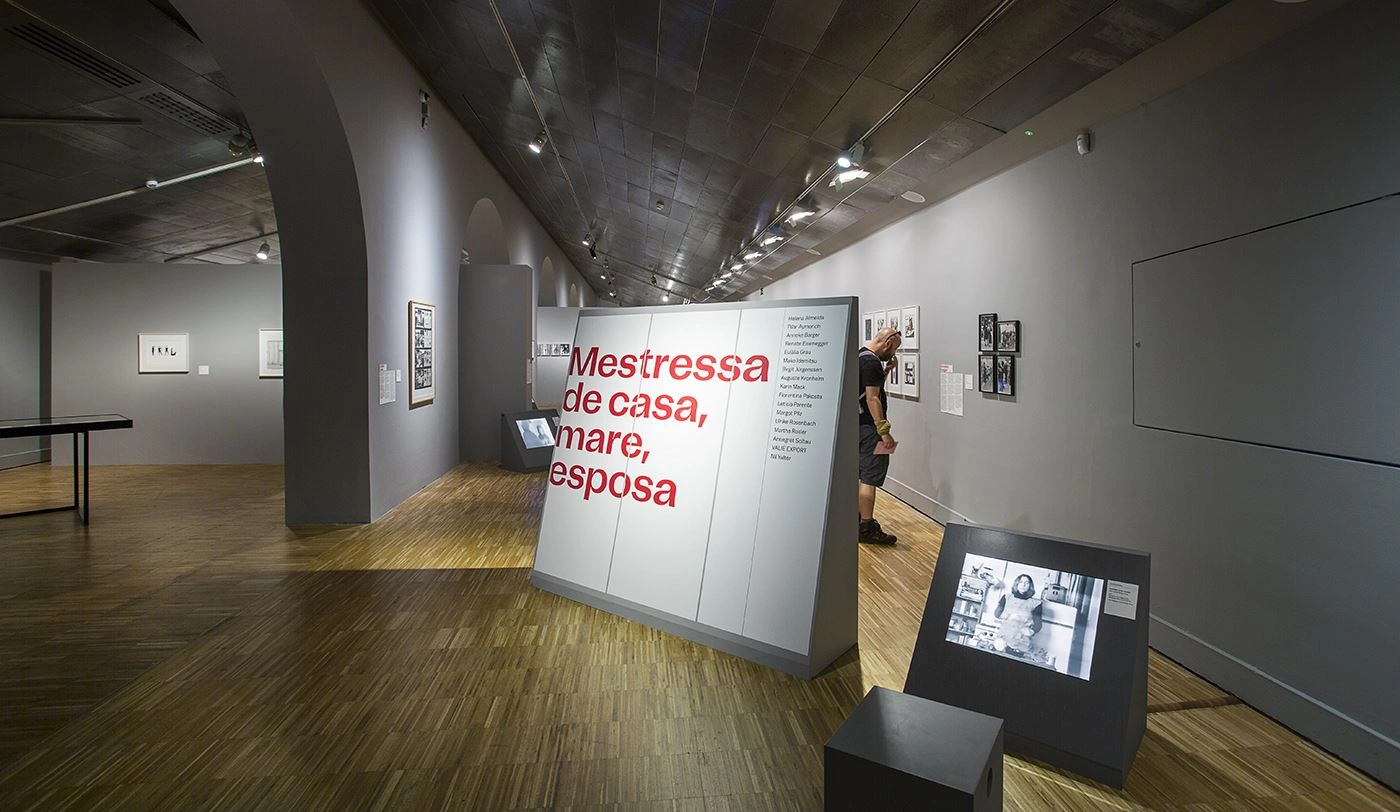 View of a modern exhibition hall featuring a large illuminated exhibit displaying the words "Mestressa de casa, mare, esposa", with several framed pictures hanging on the wall in the background and black seating furniture in the foreground.