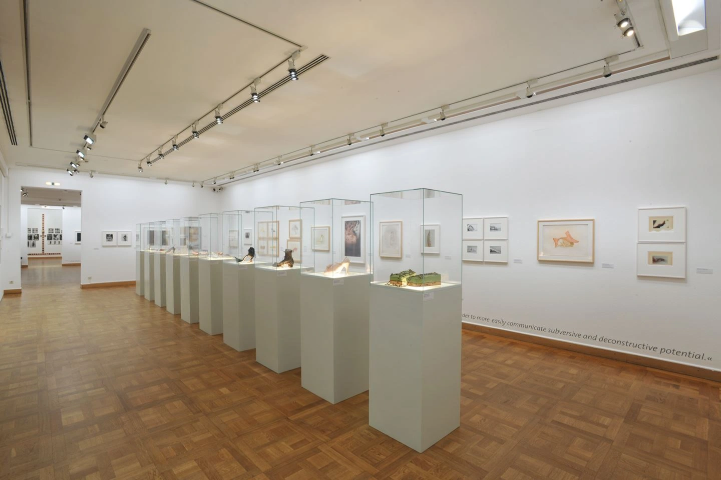 Art gallery room with bright parquet flooring and white walls adorned with various framed artworks. In the foreground, display cases with exhibits are lined up, with spotlights above providing illumination for the objects.