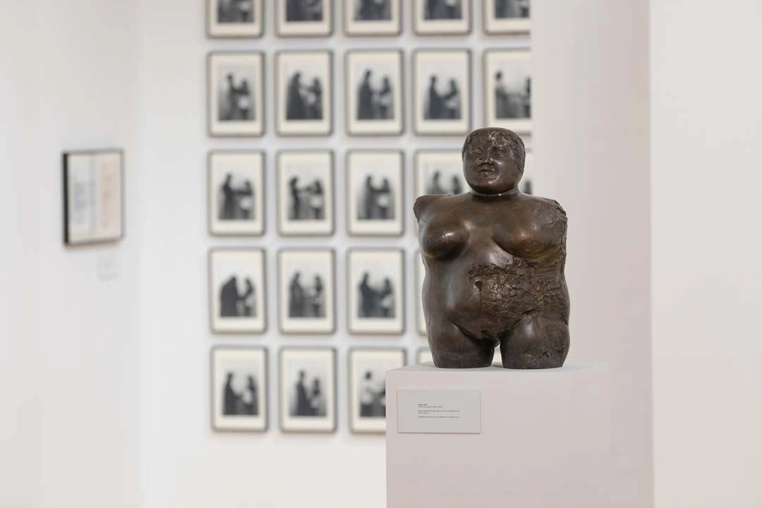 Sculpture of a human torso on a pedestal in the foreground, with a wall of framed black-and-white photographs in the background at an art exhibition.