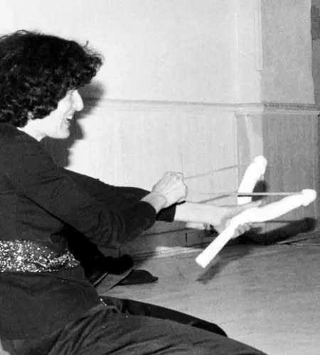 Eine Frau kniet mit einer Steinschleuder vor je einer nackten männlichen und weiblichen Aufblaspuppe und zielt auf die beiden.