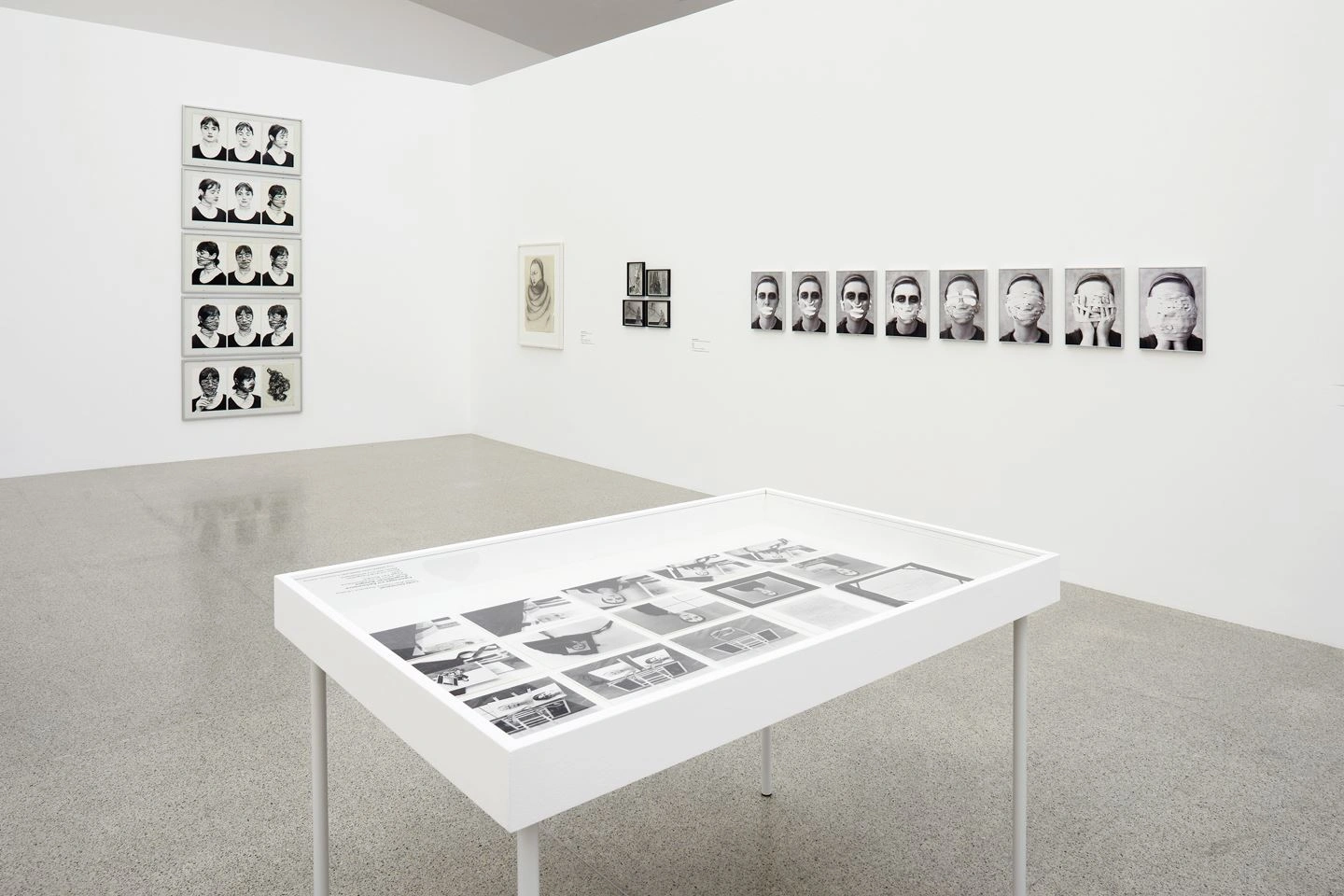 In a bright exhibition room, black and white portrait photographs are hung on two walls. In a display case in the middle of the room, pictures are also exhibited in black and white.