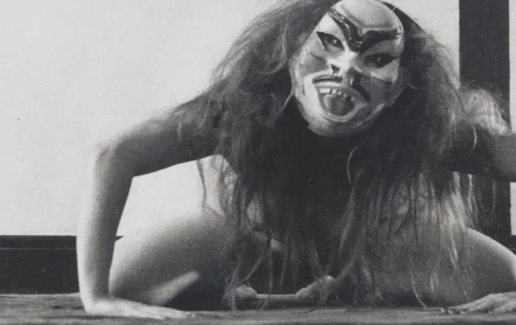 The picture shows the artist ORLAN. She is naked, kneeling on the floor, wearing a mask and her hair is tousled. The photo is in black and white.