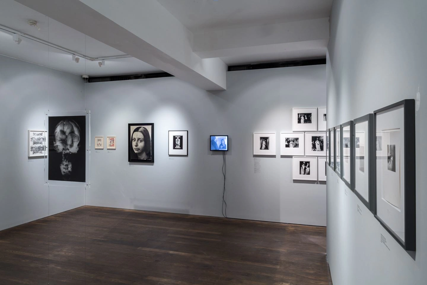 Interior view of a bright, modern art gallery with diverse black-and-white photographs and artworks on grey walls, and a blue video installation in the background, on dark polished wooden flooring.