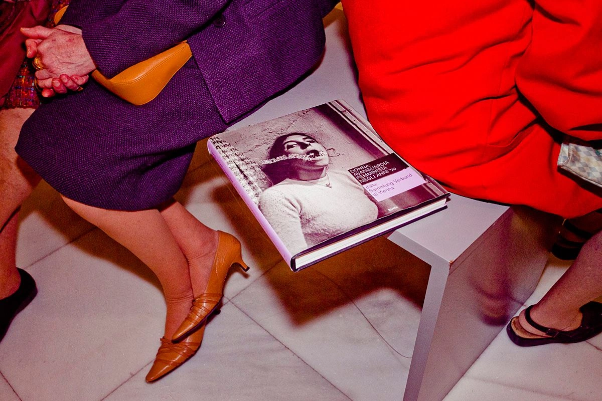 Two ladies with pointed shoes are sitting on a bench. The bound exhibition catalog lies between them.