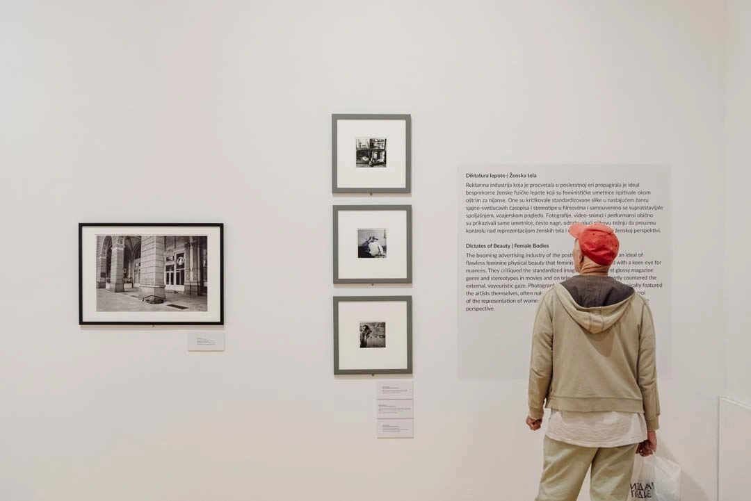 Person mit roter Mütze und beiger Jacke betrachtet eine Wand mit Schwarz-Weiß-Fotografien in unterschiedlichen Größen in einer Kunstgalerie, begleitet von Beschreibungstexten neben den Werken.