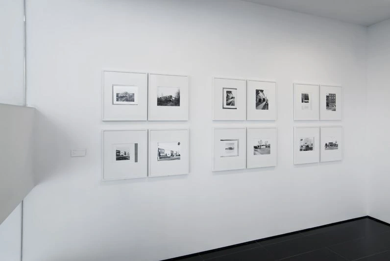 Helle Fotogallerie mit weißer Wand und dunklem Boden. Auf der weißen Wand hängen zwölf quadratische Fotografien.