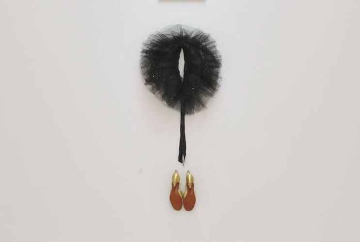 Modern art installation on a wall featuring a circular black feather object at the top, a black rod attached underneath transitioning into two golden, flame-textured shoes, against a background with an abstract brown pattern on the sides.